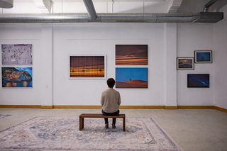 A quiet contemporary art gallery with white walls and exposed ductwork, where a person sits on a bench studying a series of framed photographs, including desert and abstract landscapes.