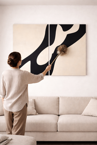 In a modern, neutral-toned living room, a person gently dusts a large abstract black-and-beige wall artwork hanging above a beige sofa.