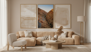 A serene living room styled in warm neutral tones, with a plush beige sofa, round wood coffee table, sculptural decor, and a triptych of framed wall art featuring abstract pieces and a winding canyon road photograph.