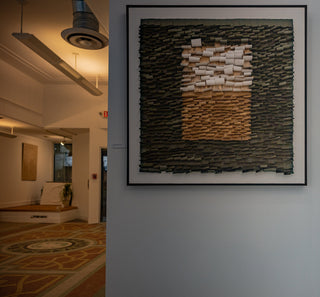 Contemporary gallery interior featuring a large framed abstract artwork composed of layered, rectangular textures in black, tan, and white, hung on a gray wall.