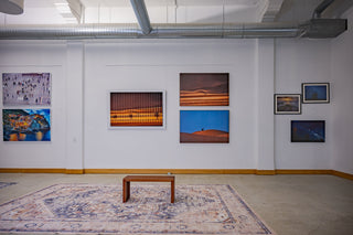 A contemporary art gallery interior with exposed ceiling ductwork, framed photographic artworks arranged along white walls, and a simple wooden bench placed on a patterned rug in the center of the room.