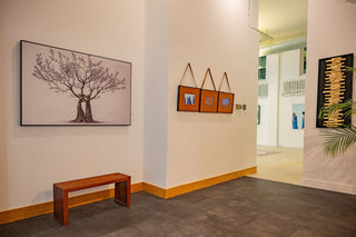 Modern art gallery interior with white walls displaying framed artworks, including a detailed tree illustration and smaller mixed-media pieces, with a wooden bench and warm lighting.