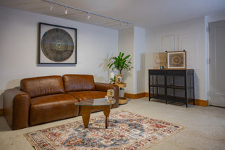 A styled gallery lounge area with a brown leather sofa on a patterned rug, a sculptural glass coffee table, potted plants, and framed abstract artwork displayed on white walls.